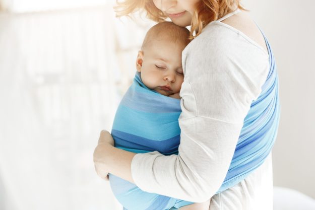 Tiny newborn child close eyes and having good sleep in baby sling feeling protection from his beautiful young mother. Family, lifestyle concept.